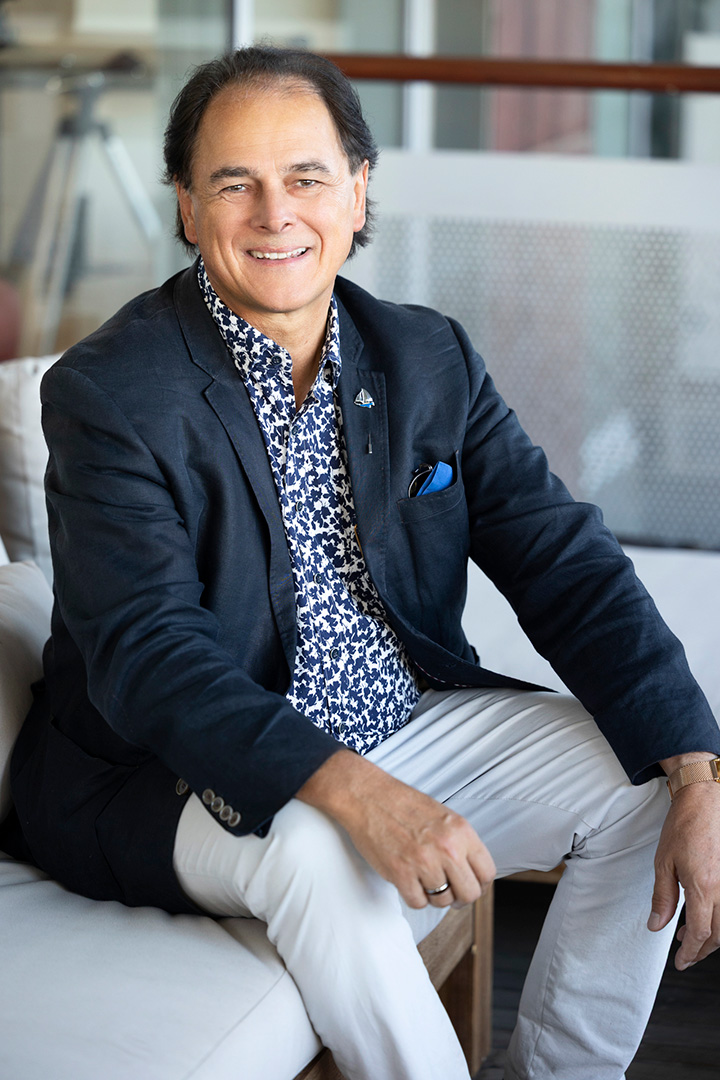 A man wearing a dark blazer, patterned shirt, and light pants sits smiling on a light-colored couch in a modern indoor setting.