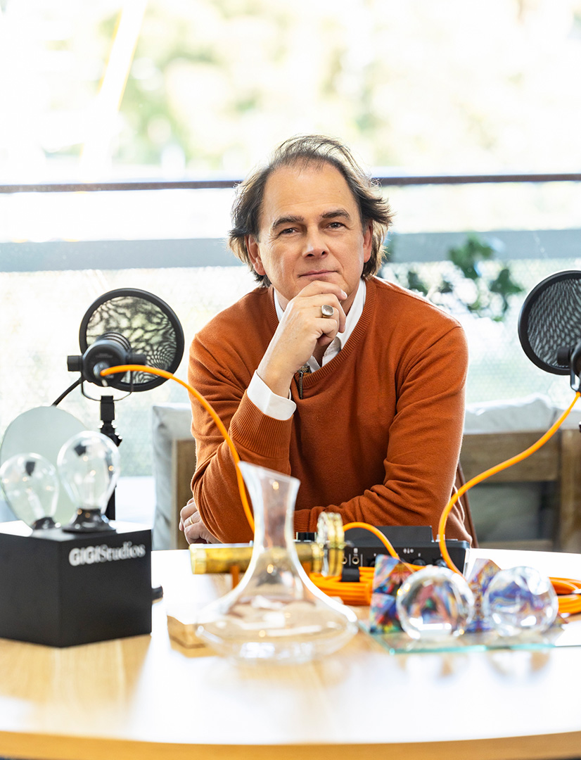 A man in an orange sweater sits at a table with a thoughtful expression. On the table are microphones, glass objects, and scientific equipment, with a bright background and greenery outside the window.