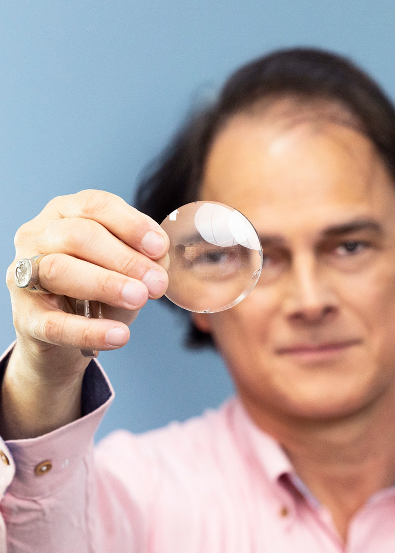A man in a pink shirt holds a clear contact lens up to the camera, partially obscuring his face and one eye, against a blue background.