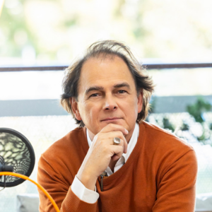 A man with shoulder-length brown hair, wearing an orange sweater over a white shirt, rests his chin on his hand and looks thoughtfully at the camera. There is a microphone in the foreground.